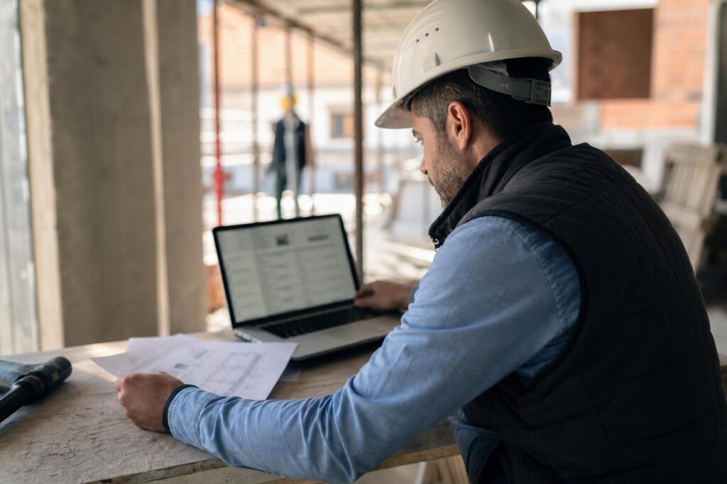 Un professionnel du BTP compare des devis de fournisseurs de matériaux sur un ordinateur portable, sur un chantier.